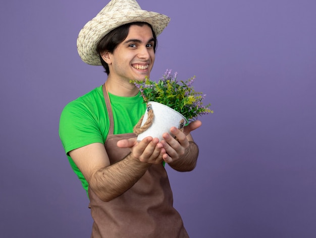 Glimlachende jonge mannelijke tuinman die in eenvormig het tuinieren hoed draagt die bloem in bloempot standhouden