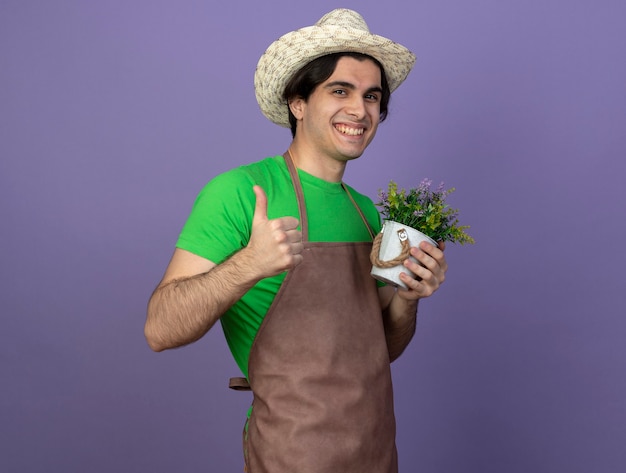 Glimlachende jonge mannelijke tuinman in eenvormig die het tuinieren de bloem van de hoedenholding in bloempot dragen die duim tonen