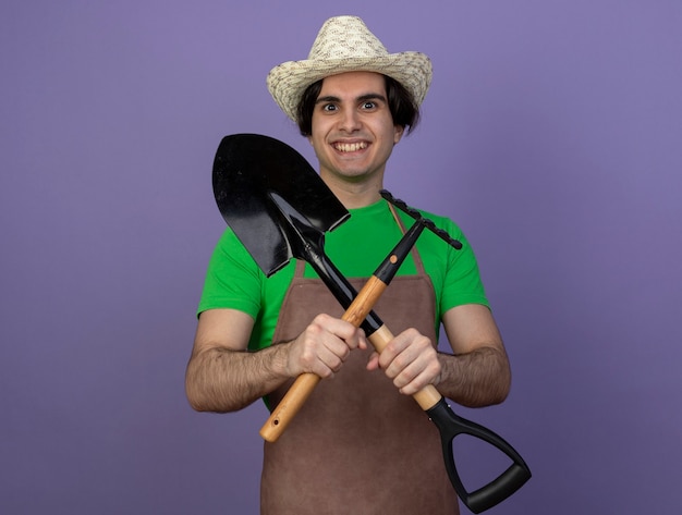Glimlachende jonge mannelijke tuinman in eenvormig die het tuinieren hoed dragen en hark met schop kruisen