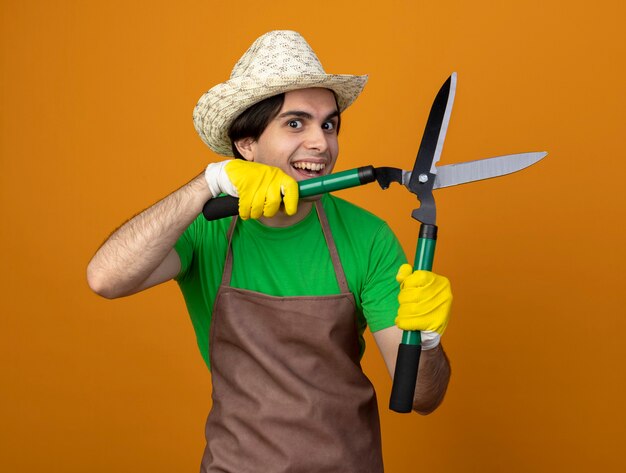 Glimlachende jonge mannelijke tuinman in uniform die tuinieren hoed met handschoenen draagt die tondeuse houden die op oranje wordt geïsoleerd