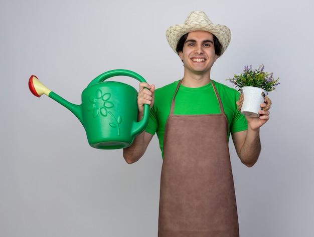 Glimlachende jonge mannelijke tuinman in uniform dragen tuinieren hoed met bloem in bloempot met gieter
