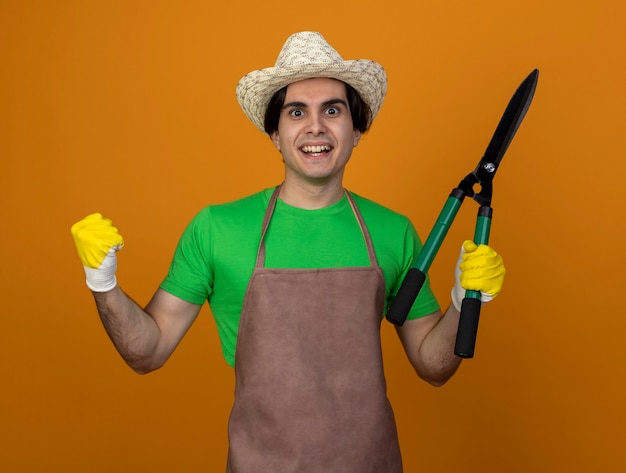 Glimlachende jonge mannelijke tuinman in uniform dragen tuinieren hoed met handschoenen met tondeuse tonen ja gebaar geïsoleerd op oranje