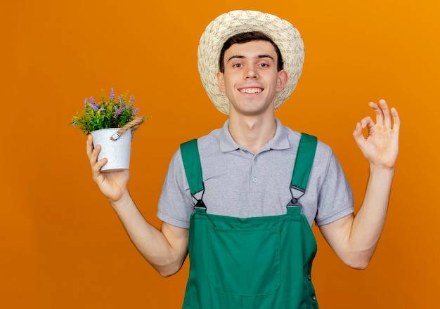 Glimlachende jonge mannelijke tuinman met tuinhoed houdt bloempot vast en gebaren ok