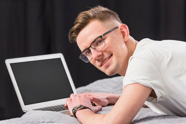 Glimlachende jonge mens die op bed liggen die weg met laptop kijken