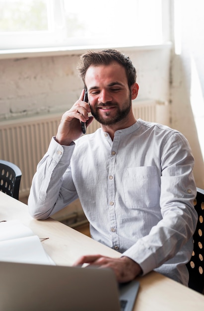 Glimlachende jonge mens die op cellphone op het werk spreekt