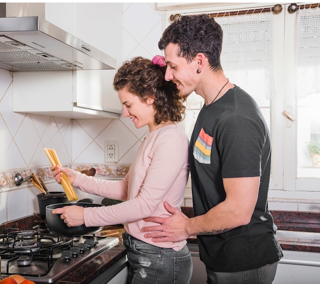 Glimlachende jonge mens die zich achter haar vrouw bevindt die spaghetti thuis voorbereidt