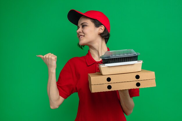 Glimlachende jonge, mooie bezorger die voedselcontainers vasthoudt met verpakkingen op pizzadozen die naar de zijkant kijken en wijzen