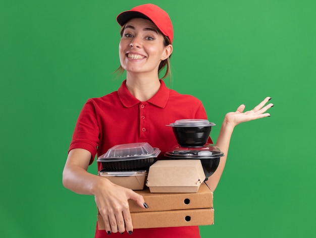 Glimlachende jonge mooie bezorger in uniform houdt papieren voedselpakketten en containers op pizzadozen en houdt de hand open geïsoleerd op groene muur