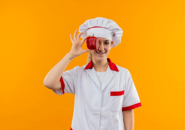 Glimlachende jonge mooie kok in eenvormige chef-kok met tandsteunen die peper op oog zetten dat op oranje ruimte wordt geïsoleerd