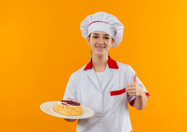 Glimlachende jonge mooie kok in eenvormige chef-kok met tandsteunen die plaat van cake houden en duim tonen die omhoog op oranje ruimte wordt geïsoleerd