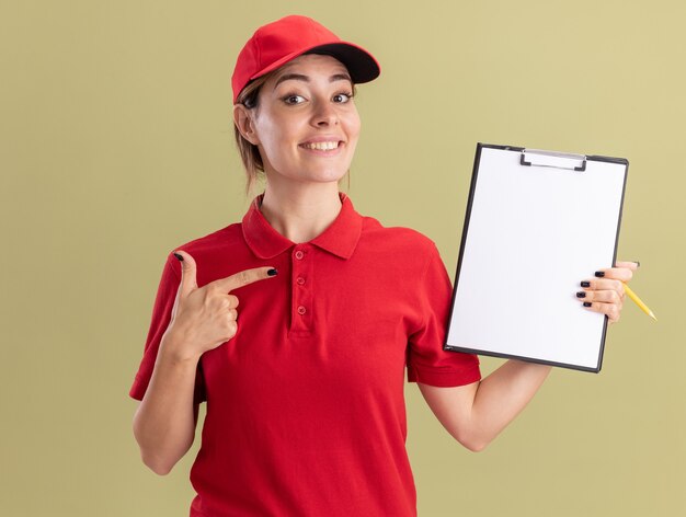 Glimlachende jonge mooie leveringsvrouw in eenvormige ruimen en punten op klembord dat op olijfgroene muur wordt geïsoleerd