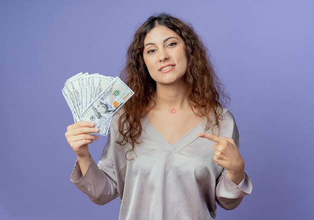 Glimlachende jonge mooie meisjesholding en punten op contant geld dat op blauwe muur wordt geïsoleerd