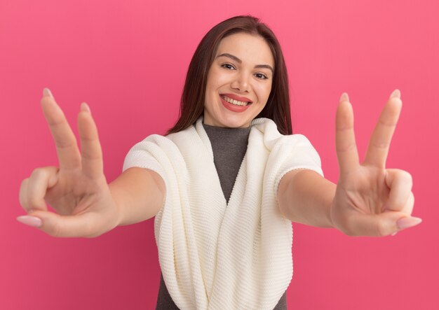 Glimlachende jonge mooie vrouw die haar handen uitstrekt naar camera die vredesteken doet geïsoleerd op roze muur