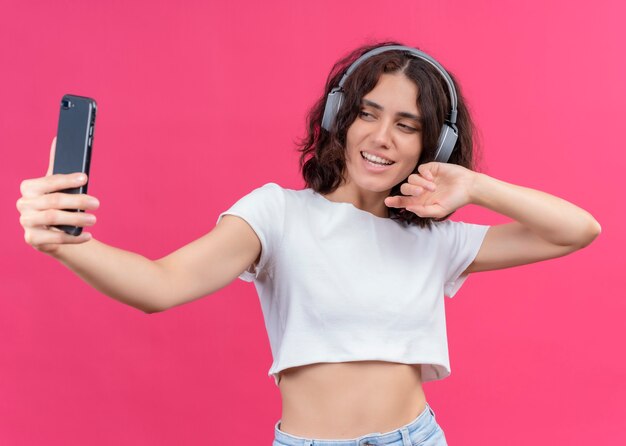 Glimlachende jonge mooie vrouw die hoofdtelefoons draagt en mobiele telefoon op roze muur houdt