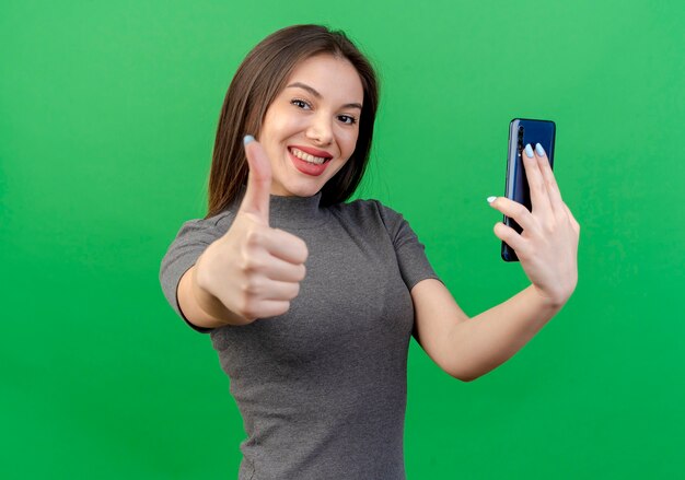 Glimlachende jonge mooie vrouw die mobiele telefoon houdt en duim toont op camera die op groene achtergrond wordt geïsoleerd