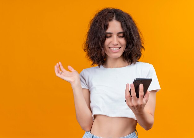 Glimlachende jonge mooie vrouw die mobiele telefoon houdt en hand op geïsoleerde oranje muur met exemplaarruimte opheft