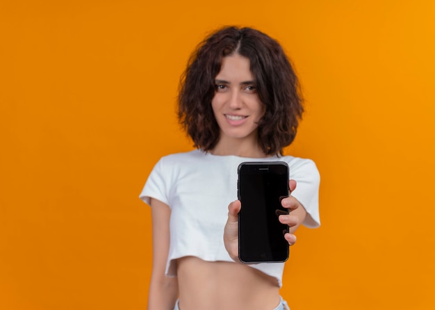 Glimlachende jonge mooie vrouw die mobiele telefoon houdt en het op geïsoleerde oranje muur met exemplaarruimte uitrekt