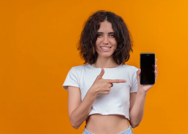 Glimlachende jonge mooie vrouw die mobiele telefoon houdt en met vinger erop richt op geïsoleerde oranje muur met exemplaarruimte