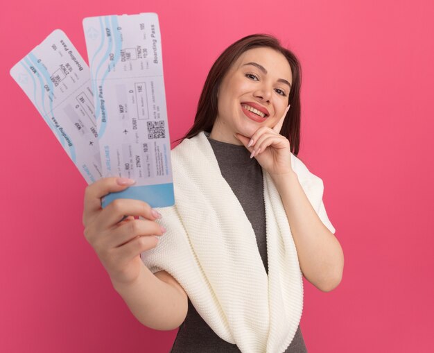 Glimlachende jonge mooie vrouw die naar de voorkant kijkt en de hand op de kin houdt en de vliegtickets naar de voorkant uitrekt, geïsoleerd op een roze muur