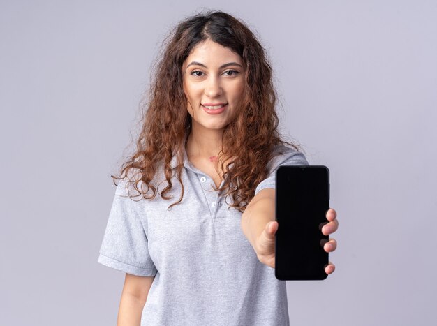 Glimlachende jonge mooie vrouw die naar de voorkant kijkt en de mobiele telefoon naar de voorkant uitrekt, geïsoleerd op een witte muur