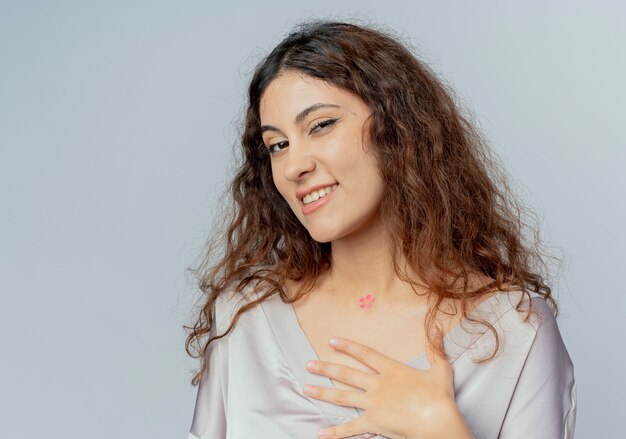 Glimlachende jonge mooie vrouwelijke beambte die hand op hart zetten dat op witte muur wordt geïsoleerd