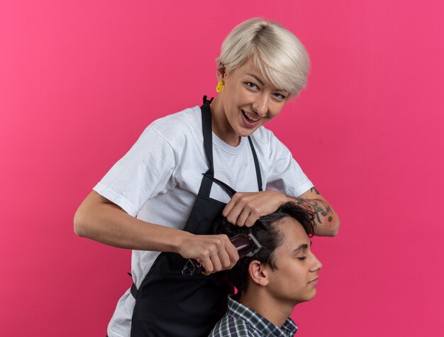 Glimlachende jonge mooie vrouwelijke kapper in uniform met kappersgereedschap en kapsel voor jongen geïsoleerd op roze achtergrond