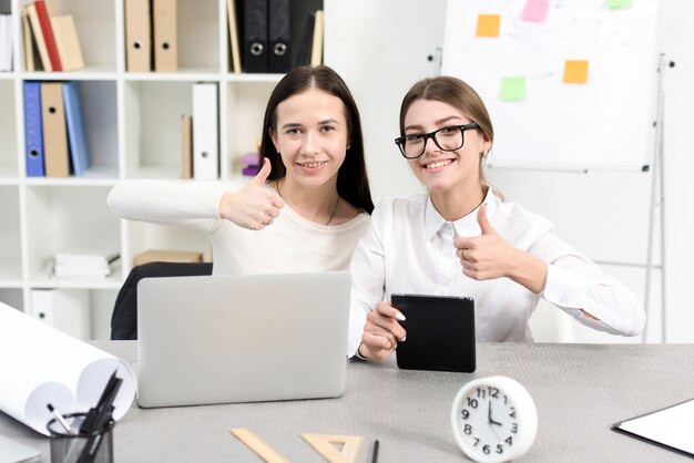 Glimlachende jonge onderneemster met laptop en digitale tablet die duim op teken op het werk tonen