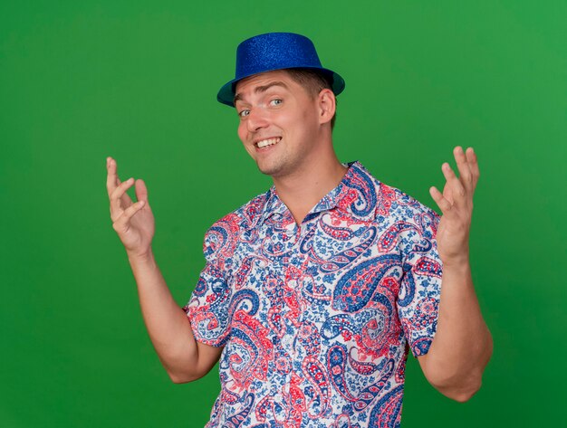 Glimlachende jonge partijkerel die blauwe hoed draagt die handen uitspreidt die op groen worden geïsoleerd