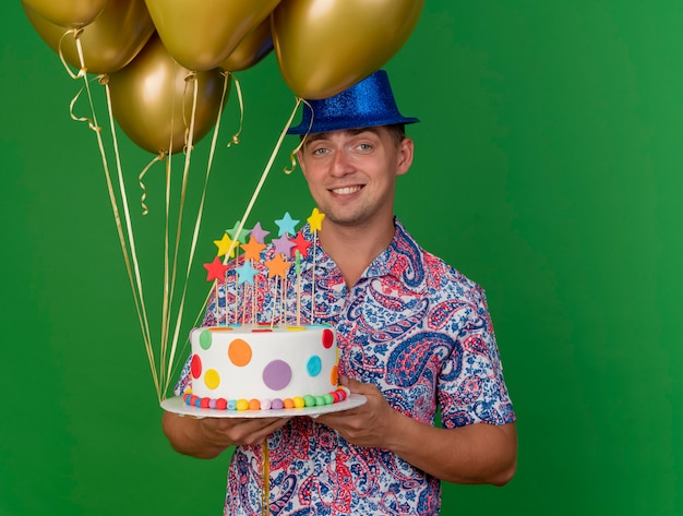 Glimlachende jonge partijkerel die de blauwe ballons van de hoedenholding met cake draagt die op groen wordt geïsoleerd