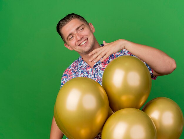 Glimlachende jonge partijkerel die kant bekijkt die kleurrijk overhemd draagt dat zich achter ballons bevindt die op groen worden geïsoleerd