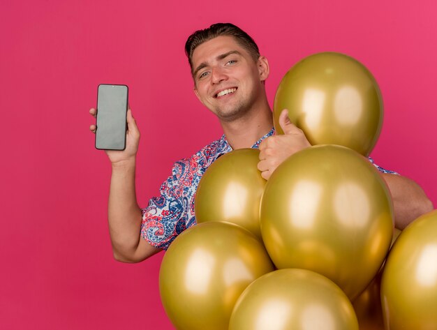 Glimlachende jonge partijkerel die kleurrijk overhemd draagt dat zich onder ballons bevindt die duim tonen die omhoog op roze wordt geïsoleerd