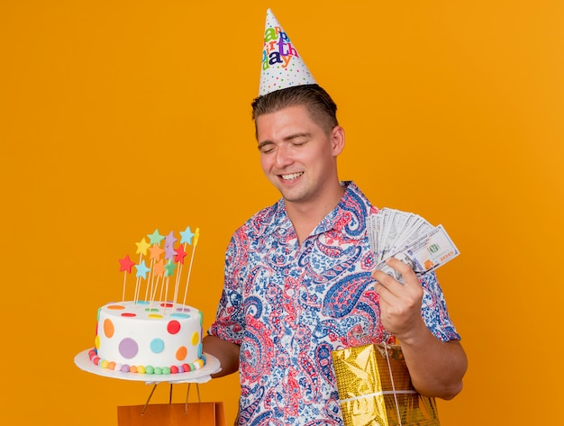 Glimlachende jonge partijkerel met gesloten ogen die verjaardag GLB dragen die cake met giften en geld houden die op oranje wordt geïsoleerd