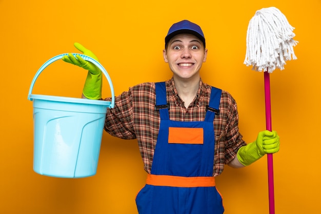 Glimlachende jonge schoonmaakster met uniform en pet met handschoenen met dweil en emmer