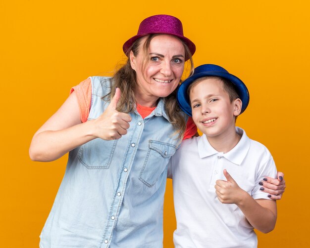 Glimlachende jonge Slavische jongen met blauwe feestmuts staande met zijn moeder met paarse feestmuts duimen omhoog geïsoleerd op oranje muur met kopieerruimte