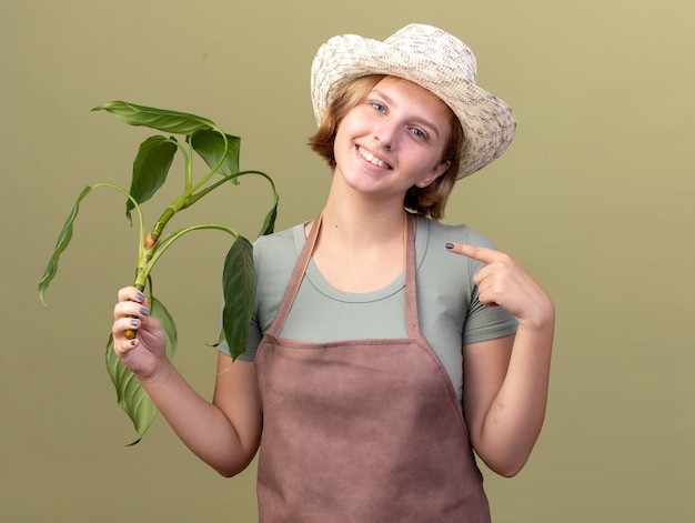 Glimlachende jonge Slavische vrouwelijke tuinman die het tuinieren hoed draagt en op plant op olijfgroen richt