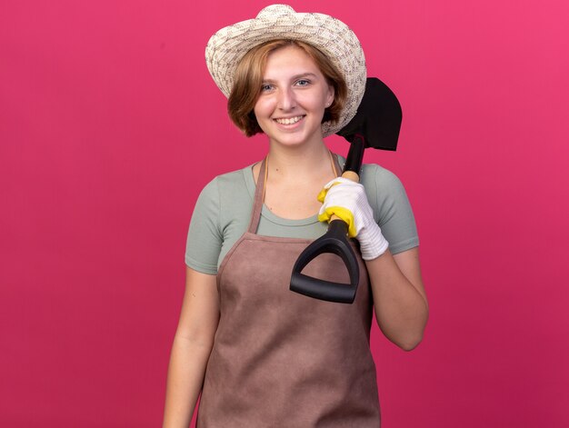 Glimlachende jonge Slavische vrouwelijke tuinman die het tuinieren hoed en handschoenen draagt die schop op schouder op roze houden