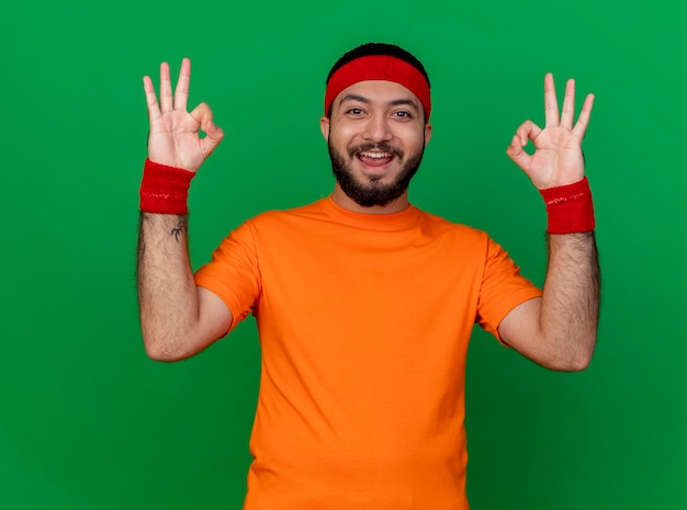 Glimlachende jonge sportieve man met hoofdband en polsbandje met ok gebaar geïsoleerd op groene achtergrond