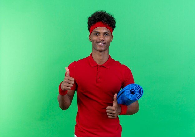 Glimlachende jonge sportieve man met hoofdband en polsbandje met yogamat