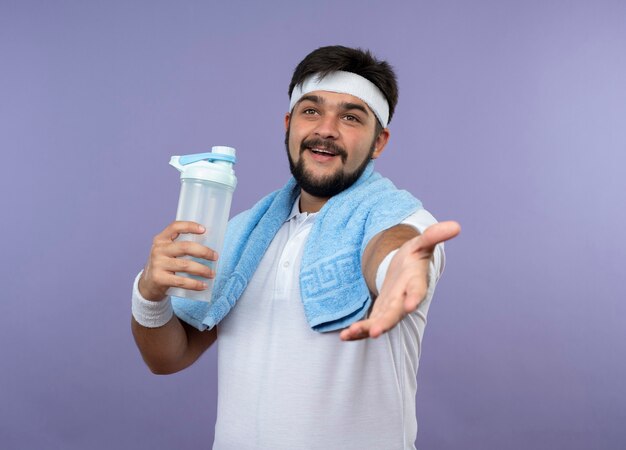 Glimlachende jonge sportieve mens die hoofdband en polsband draagt die waterfles met handdoek op schouder houdt en hand standhouden