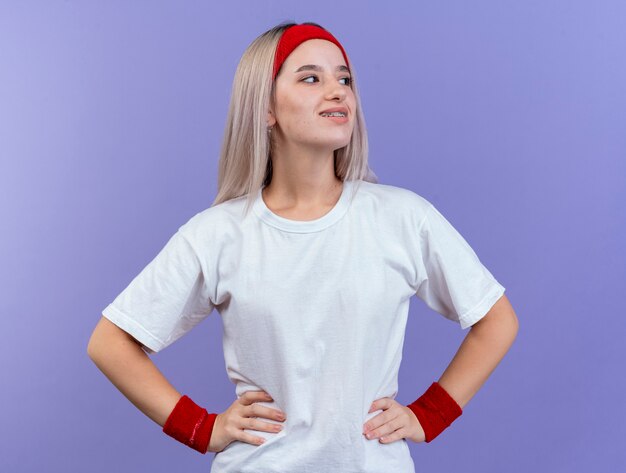 Glimlachende jonge sportieve vrouw met bretels die hoofdband en polsbandjes dragen die handen op taille zetten en kant bekijken die op purpere muur wordt geïsoleerd