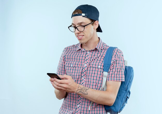 Glimlachende jonge studentenjongen die achterzak en glazen en GLB dragen die en telefoon op wit houden bekijken