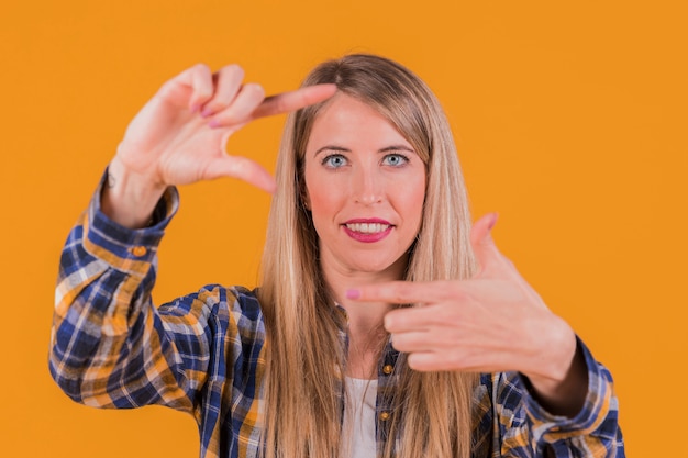 Glimlachende jonge vrouw die door handframe kijkt tegen een oranje achtergrond