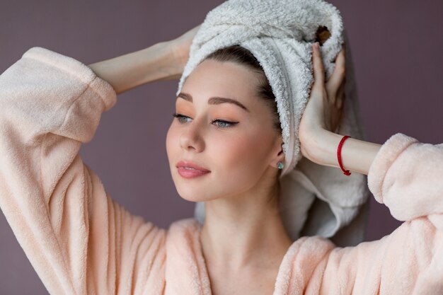 glimlachende jonge vrouw die een badjas en een handdoek op haar hoofd draagt en op roze staat