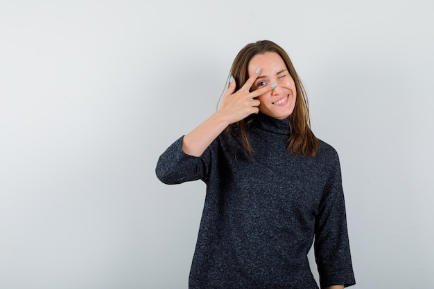 Glimlachende jonge vrouw die een teken van de overwinningshand in haar linkeroog op witte achtergrond houdt