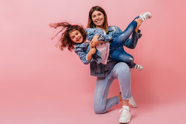 Glimlachende jonge vrouw die met dochter speelt. Studio shot van gelukkige moeder en preteen kind in denim kleding.