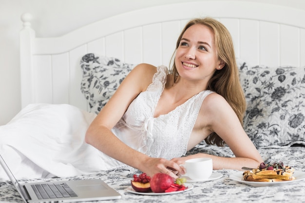 Glimlachende jonge vrouw die op bed ligt dat van het ochtendontbijt geniet