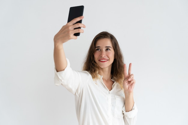 Glimlachende jonge vrouw die selfie foto neemt en overwinningsteken toont.