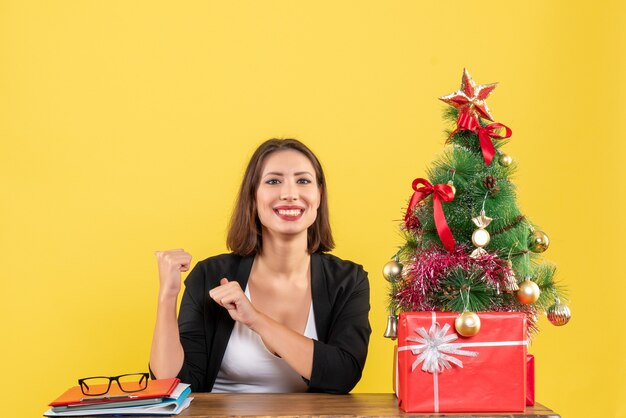 Glimlachende jonge vrouw die van haar succes in kostuum geniet dichtbij verfraaide kerstboom op kantoor op geel