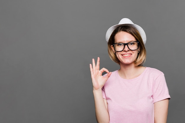 Glimlachende jonge vrouw die witte hoed draagt die ok teken toont tegen grijze achtergrond