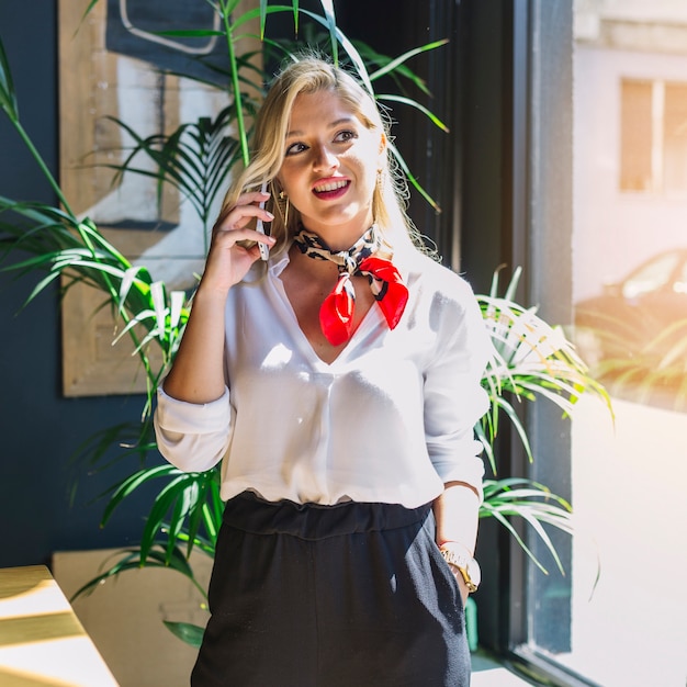 Glimlachende jonge vrouw die zich dichtbij het venster bevindt dat op mobiele telefoon spreekt